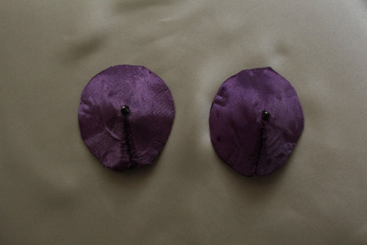 A pair of Round Purple satin pasties on a cream background shot from above.